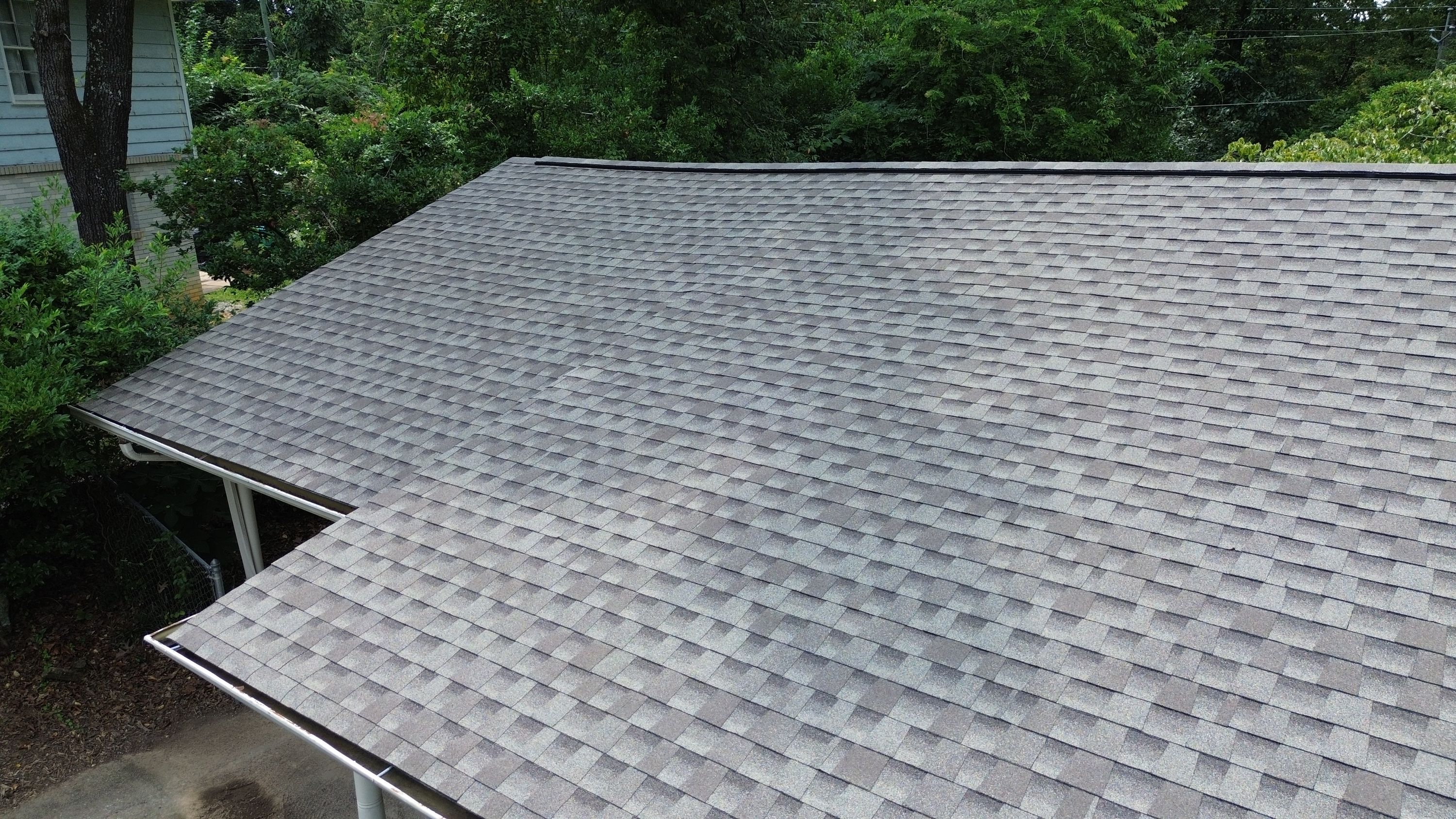 Freshly laid architectural shingles run evenly up to the ridge cap against a backdrop of dense green trees.