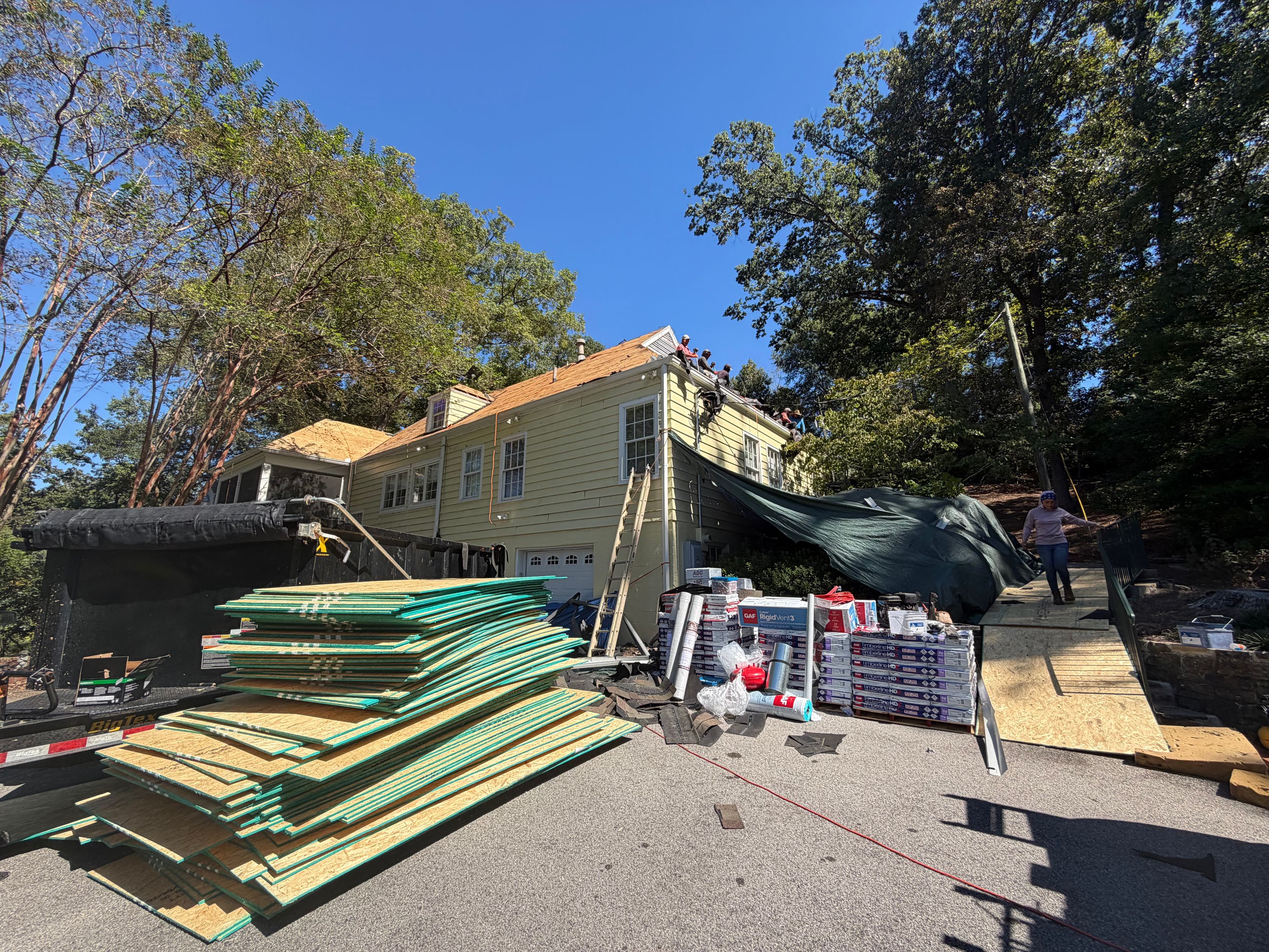 Stacked shingle roofing supplies and plywood line the driveway of a home undergoing a roof replacement.
