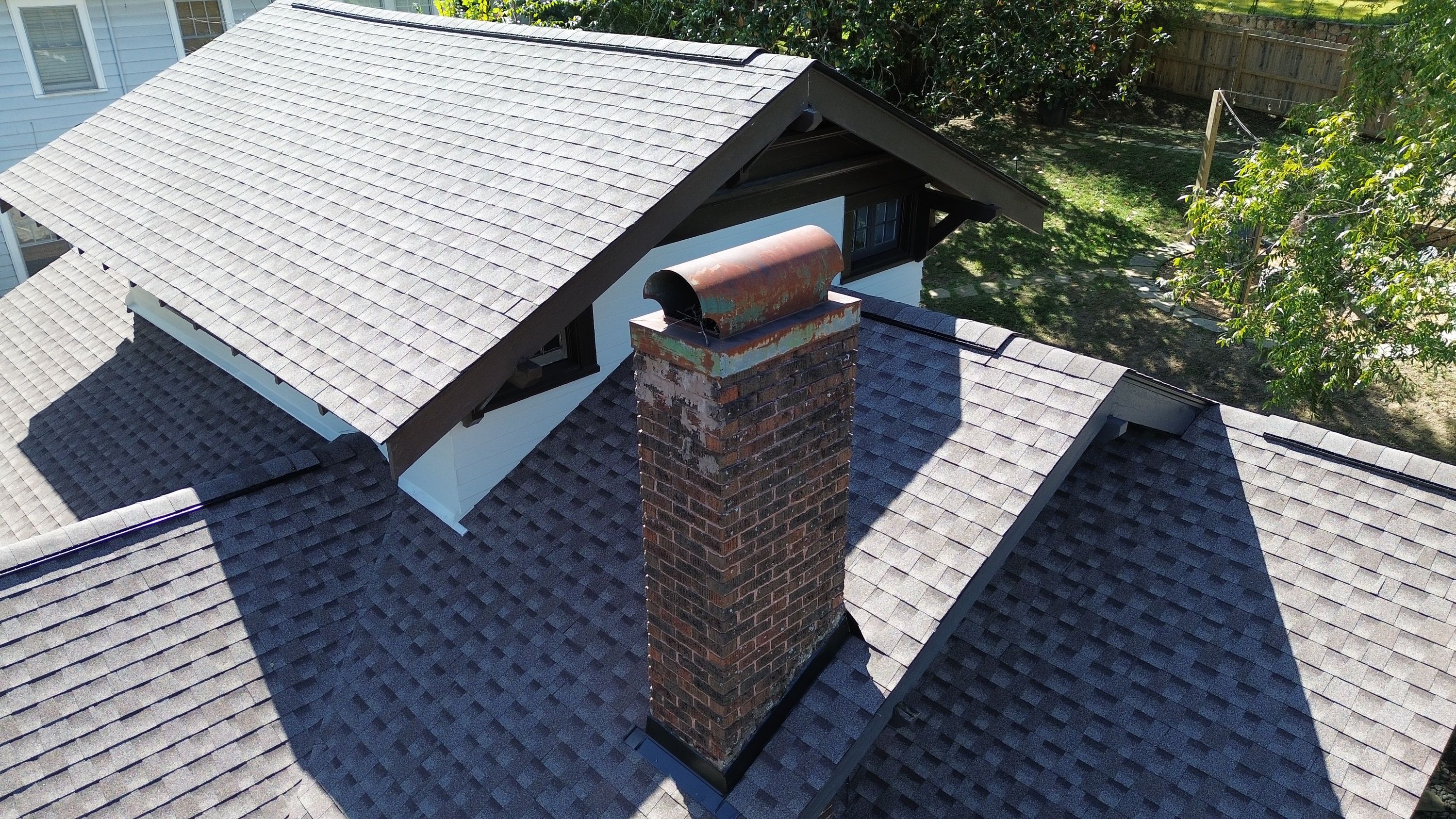 Brick chimney with black base flashing rising through a lower shingle roof slope in front of a white gable.