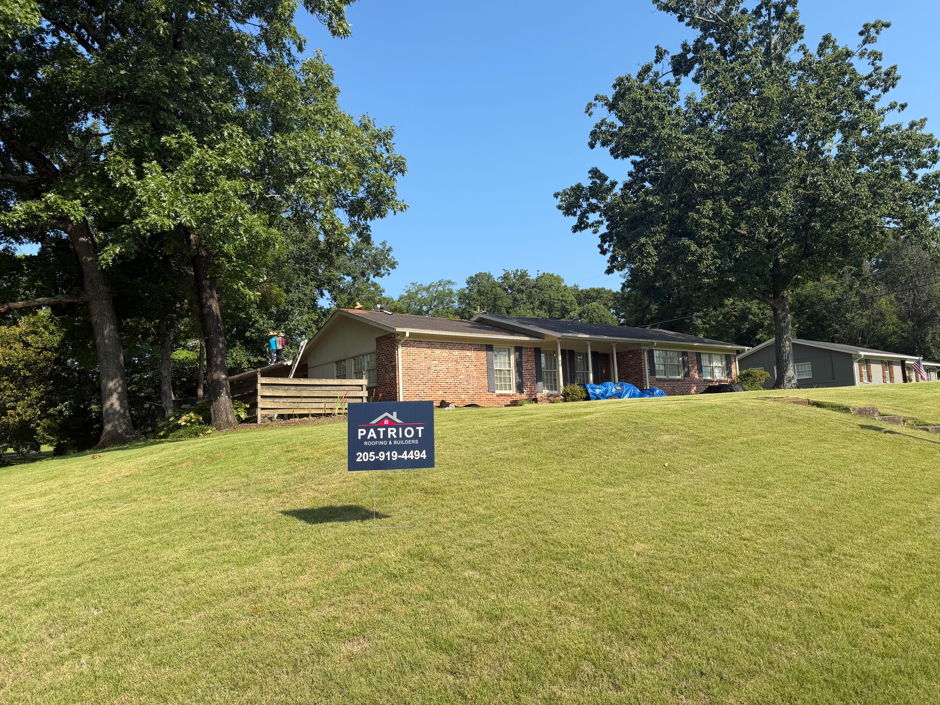 A red brick home on Columbiana Road undergoes a roof replacement, shown with shingles stripped on a sunny day.